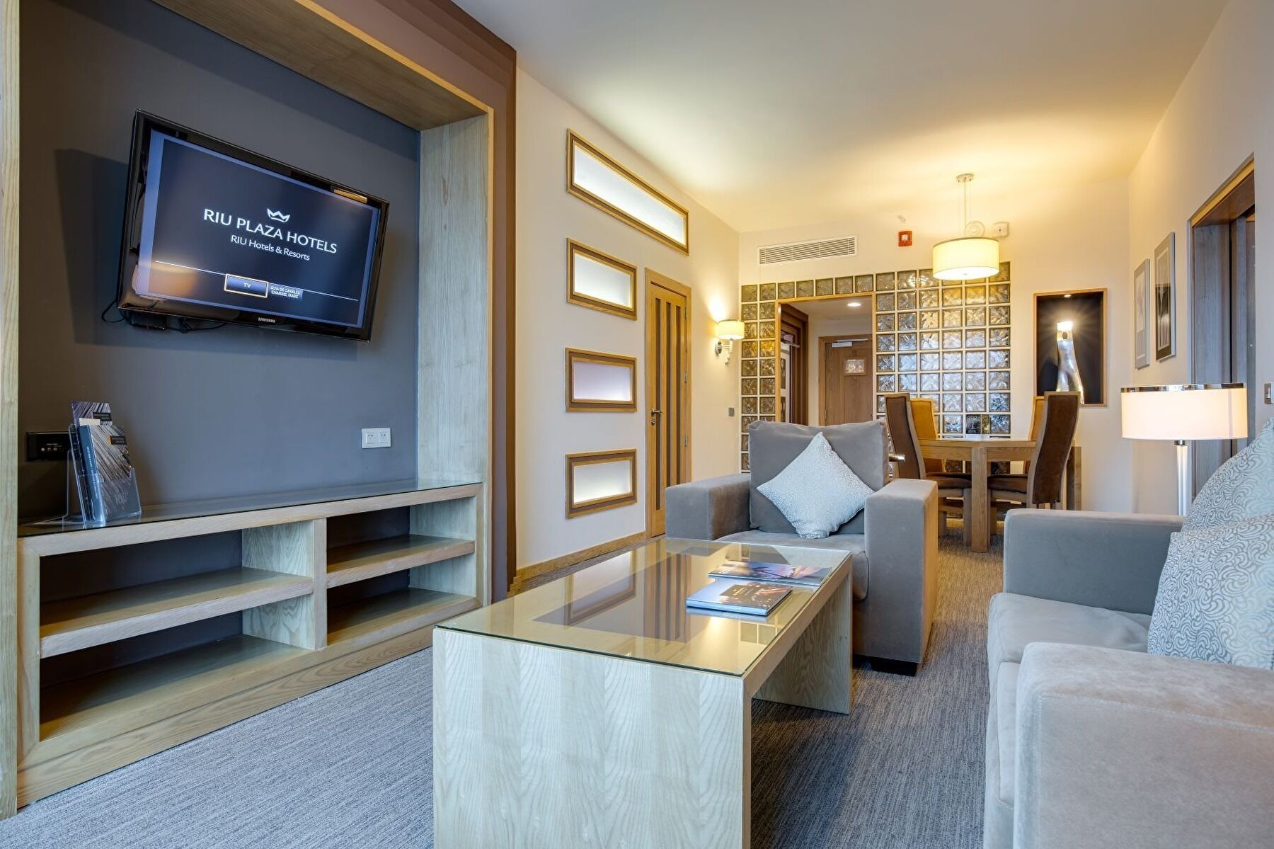 A modern hotel room with a flat-screen TV, a glass coffee table, and a seating area with a sofa and armchairs.