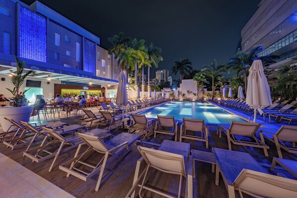 A poolside area with lounge chairs and umbrellas, a bar, and a building with a modern design.