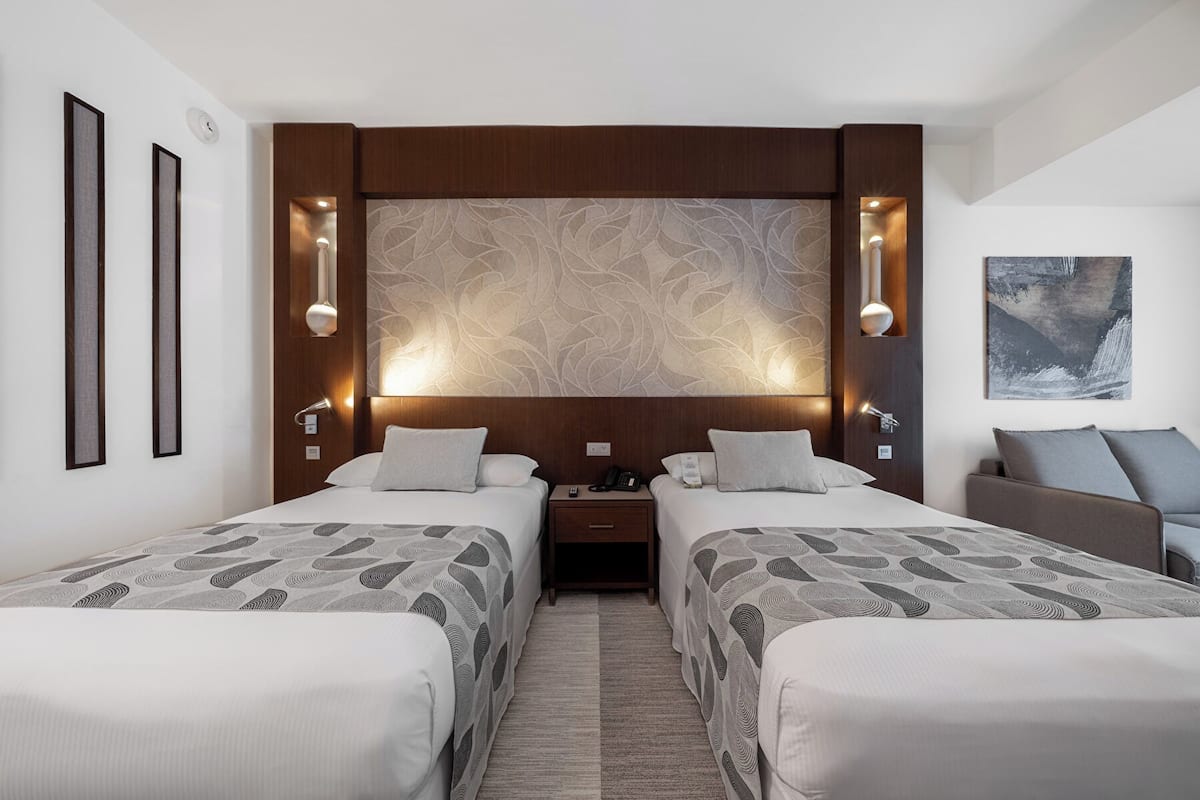 Habitación de hotel con dos camas, un cabecero decorativo grande, lámparas de noche, mesitas de noche y un cuadro en la pared.