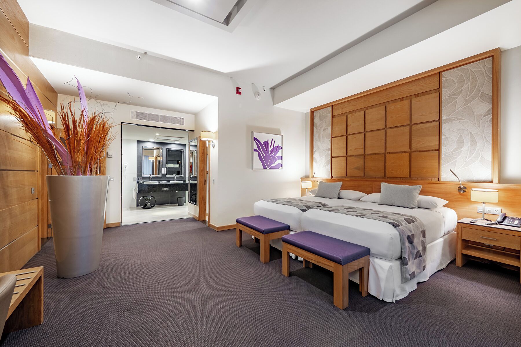 Une chambre d’hôtel moderne avec un grand lit, deux tables de chevet, une tête de lit en bois, une grande fenêtre et une salle de bain visible à travers une porte ouverte.