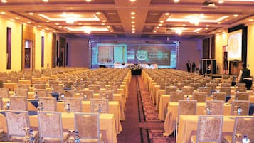 A conference room with rows of tables and chairs, a stage with a presentation screen, and a carpeted floor.