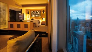 A hotel room with a bed, a TV, a desk, and a view of the cityscape through a window.