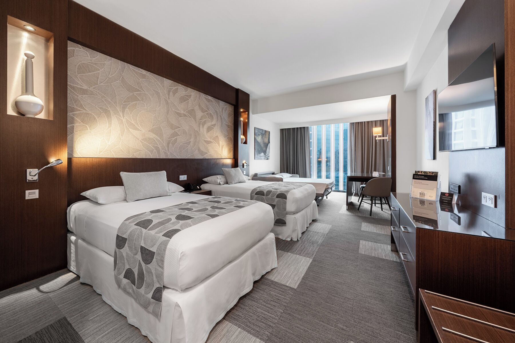 A hotel room with two beds, a desk, a chair, a TV, and a large window with curtains.