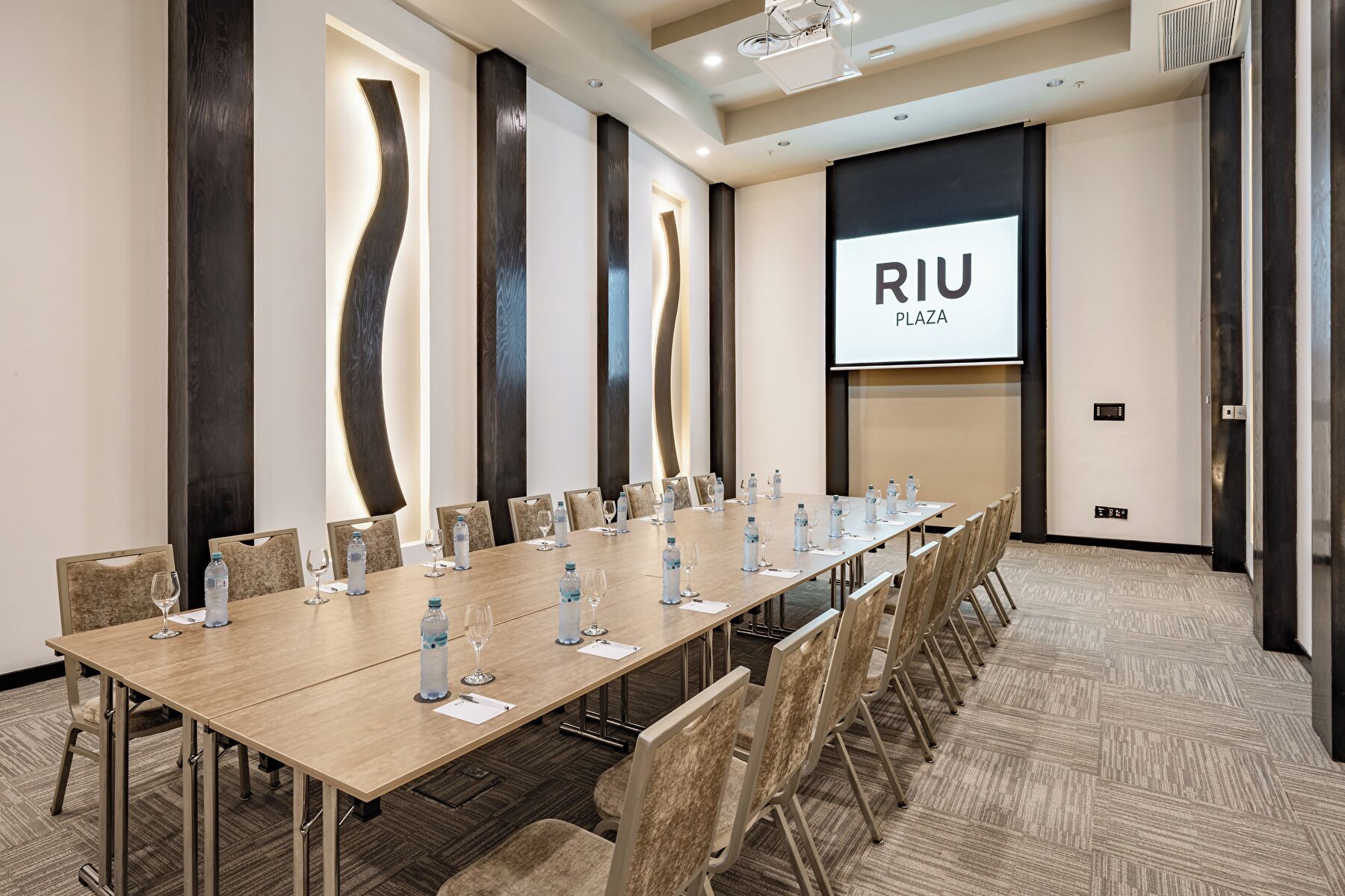 A conference room with a long table, chairs, water bottles, and a projector screen displaying "RIU PLAZA".