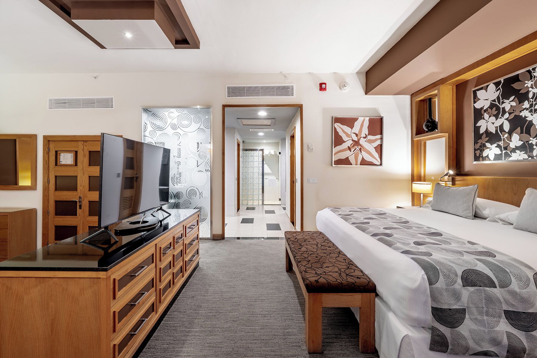 A hotel room with a bed, a desk with a flat-screen TV, a bench, and a door leading to a hallway.