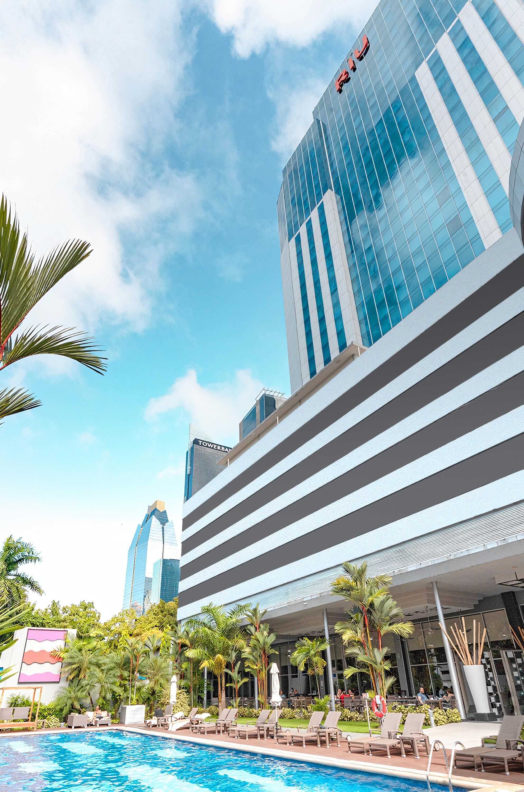 A modern hotel with a pool area, lounge chairs, and palm trees.