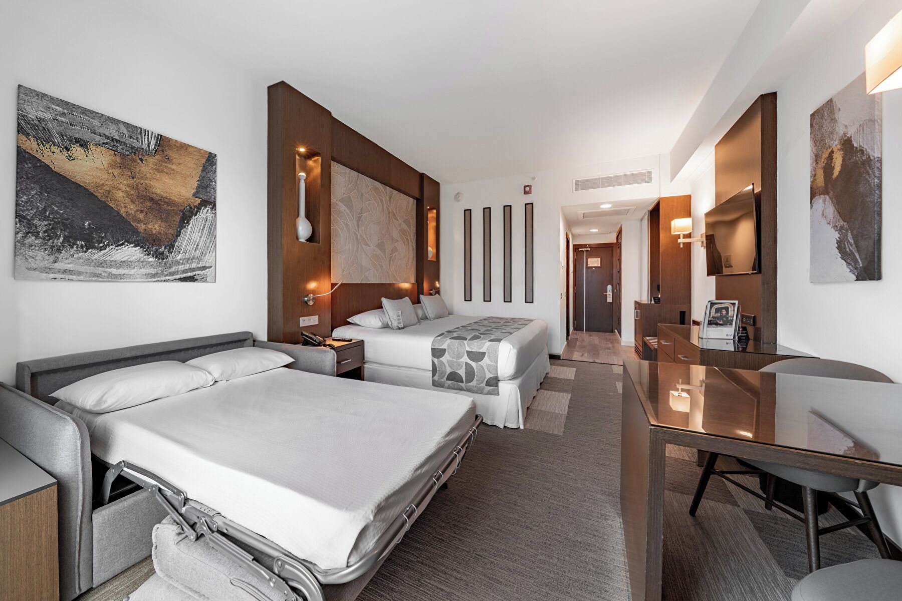 Habitación de hotel moderna con dos camas, un escritorio y un baño visible al fondo.