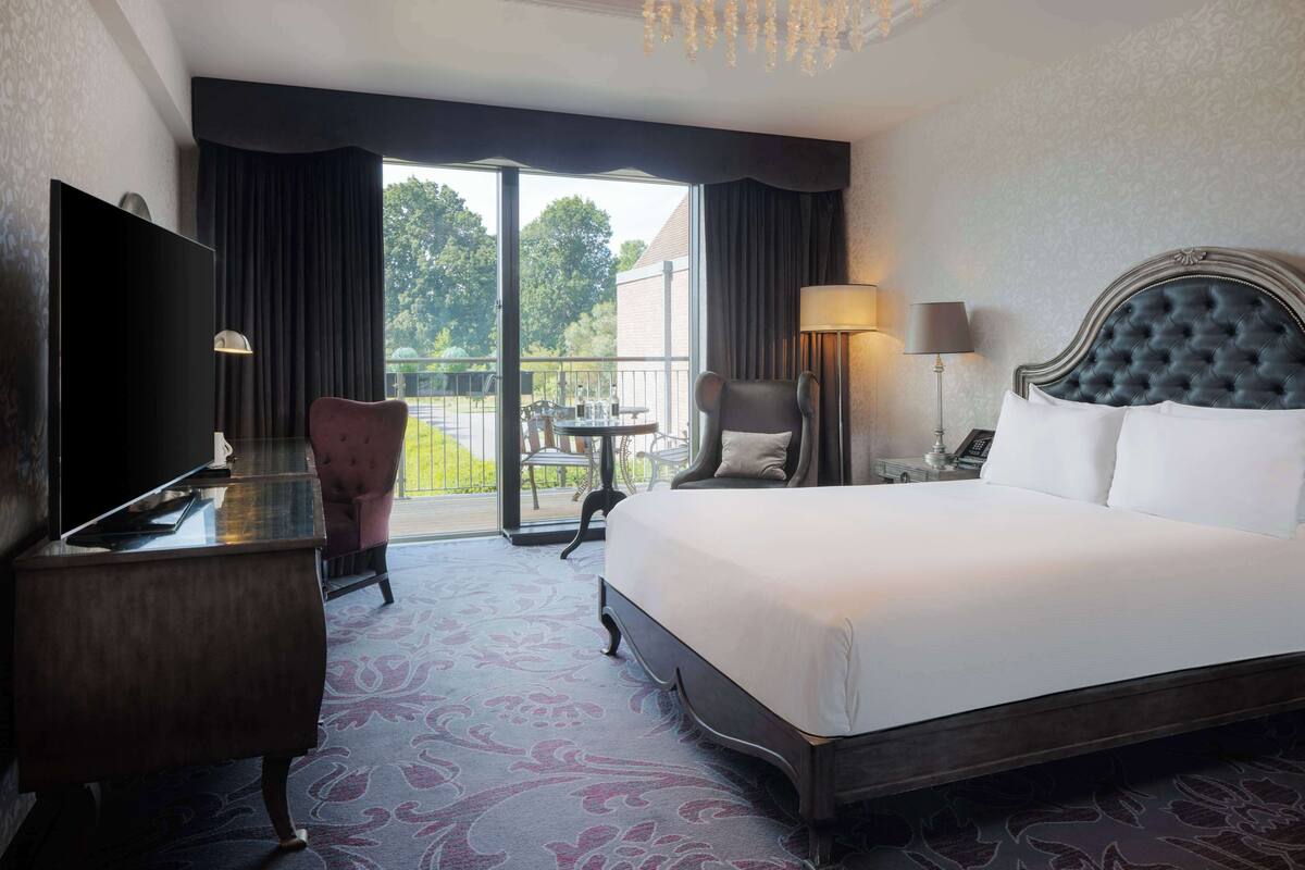 Quarto de hotel com uma cama grande, uma escrivaninha, uma cadeira, uma televisão e uma varanda com mesa e cadeiras.