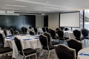 A conference room with round tables, chairs, and a projector screen. - Hilton London Syon Park (Brentford)