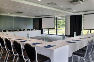 Un salón de conferencias con una larga mesa, sillas, una pantalla de proyector y vistas al exterior.