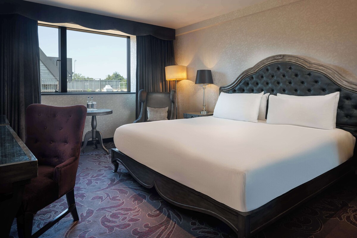 A hotel room with a large bed, a desk, a chair, a lamp, and a view of the outside.