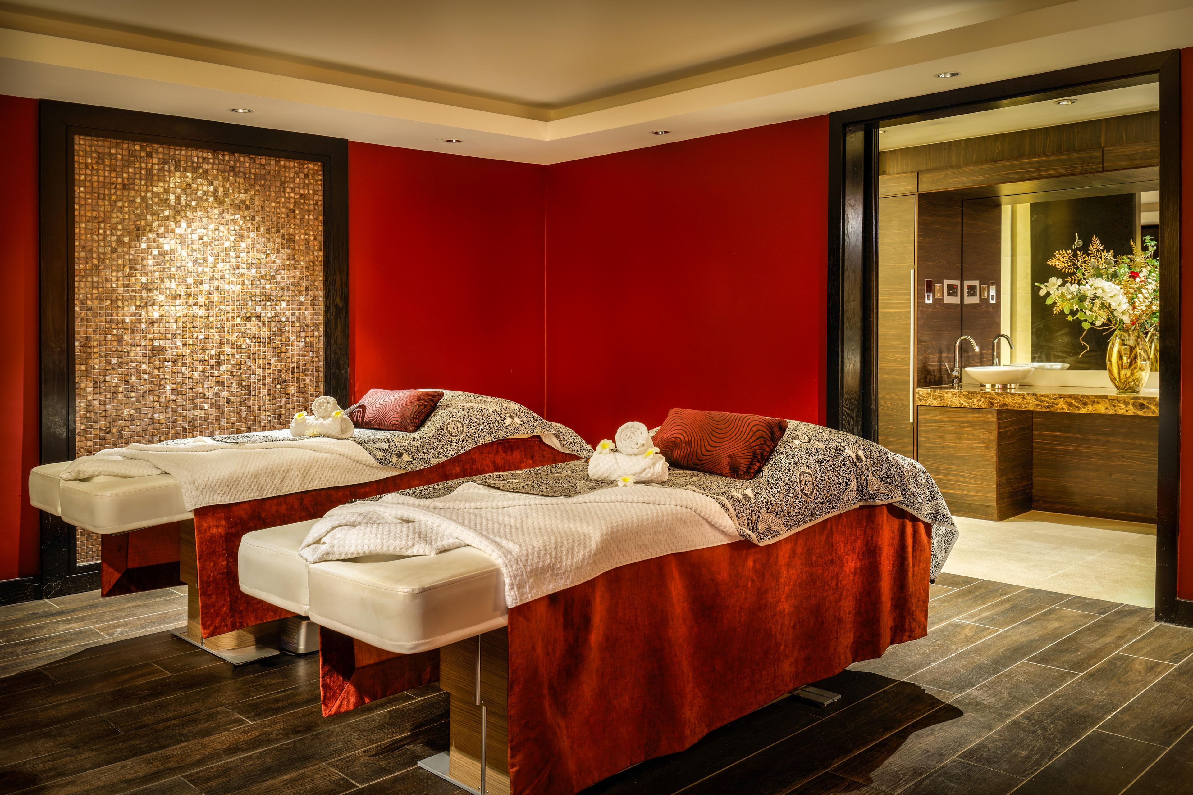 A spa room with two massage tables, a red wall, a decorative mosaic, and a mirror with a sink and flowers.