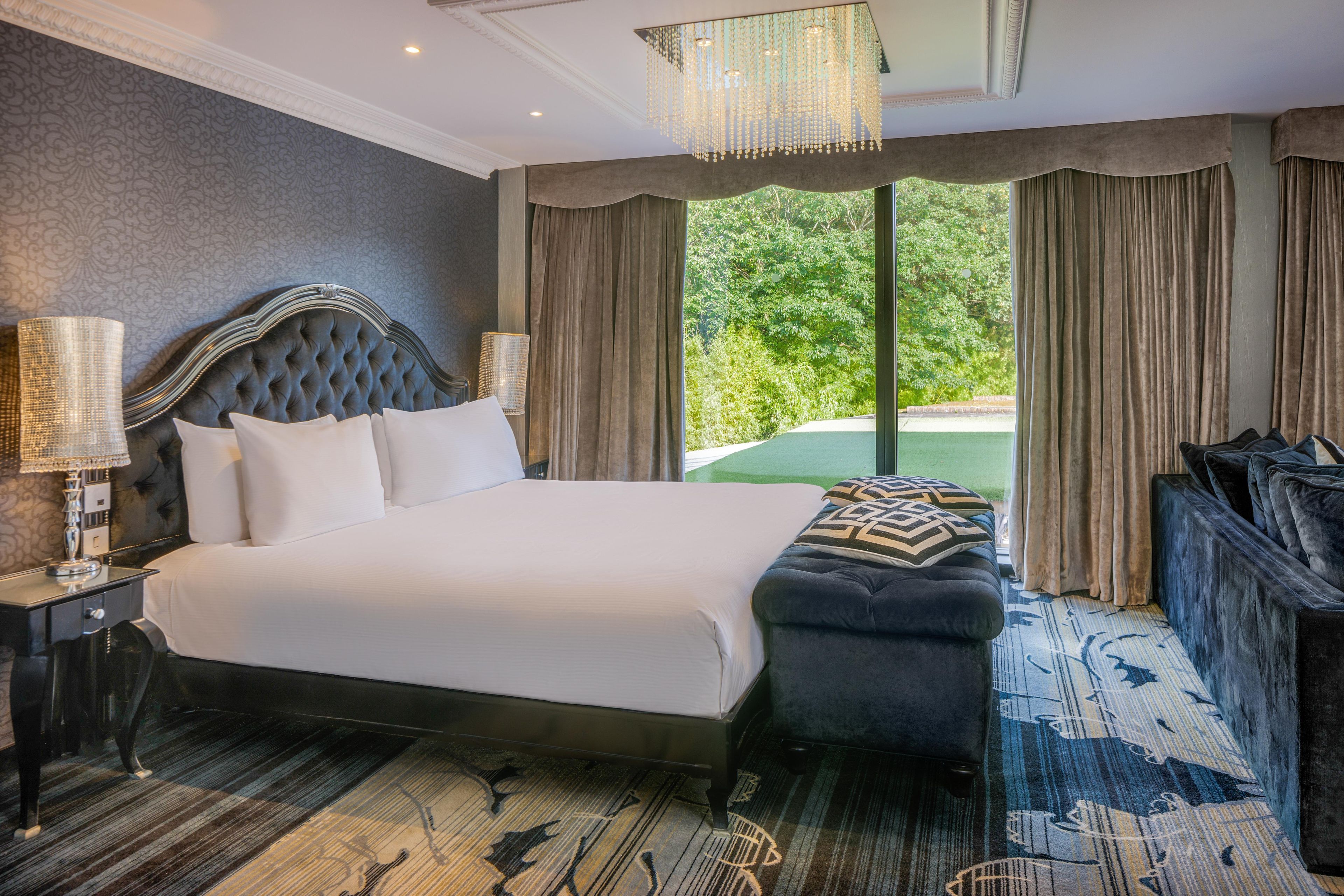 A large bed with a tufted headboard, a bedside lamp, and a bench with a patterned cushion.