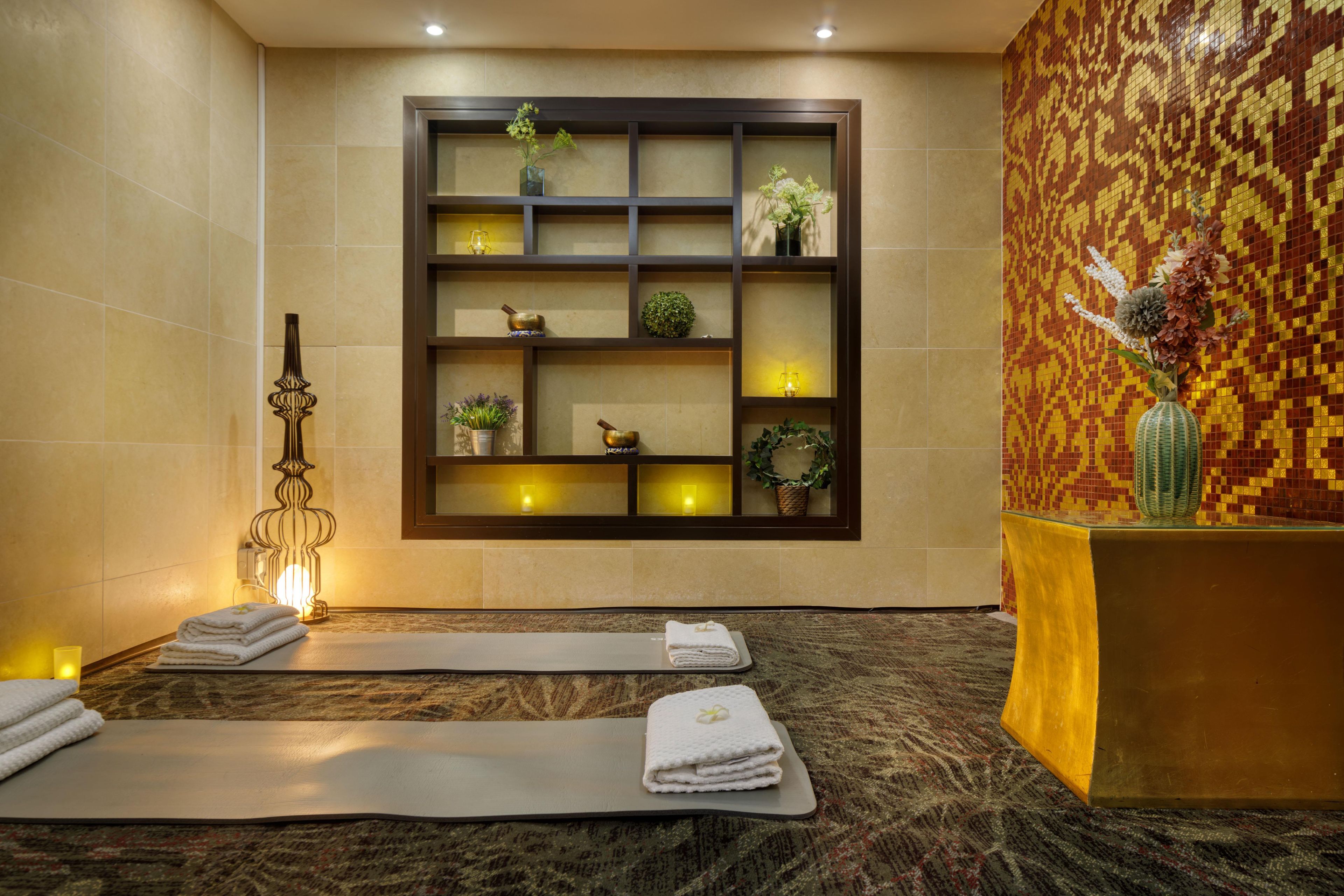 A spa room with a massage table, towels, a decorative wall, and a shelf with plants and candles.