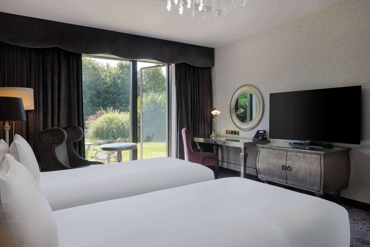 Quarto de hotel com duas camas, uma escrivaninha com televisão, uma cadeira e vista para área verde pela janela.