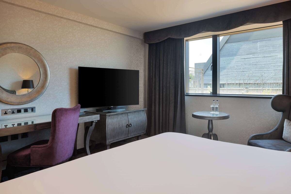 Quarto de hotel com uma cama grande, televisão de tela plana, penteadeira com espelho, mesa redonda pequena e uma cadeira.