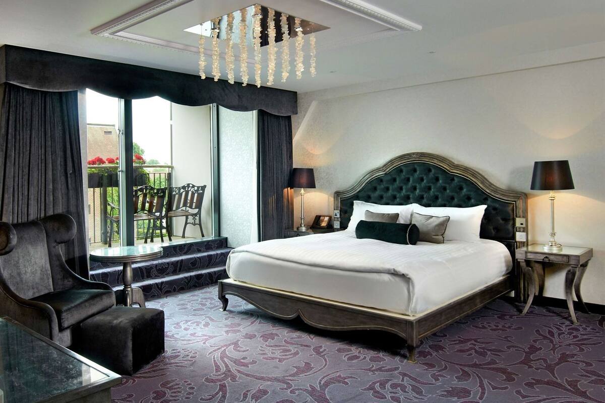Quarto de hotel com uma cama grande, um candelabro, varanda com cadeiras e vista para o exterior.