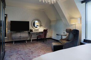 A hotel room with a flat-screen TV, a desk with a lamp, a chair, and a round mirror on the wall. - Hilton London Syon Park (Brentford)