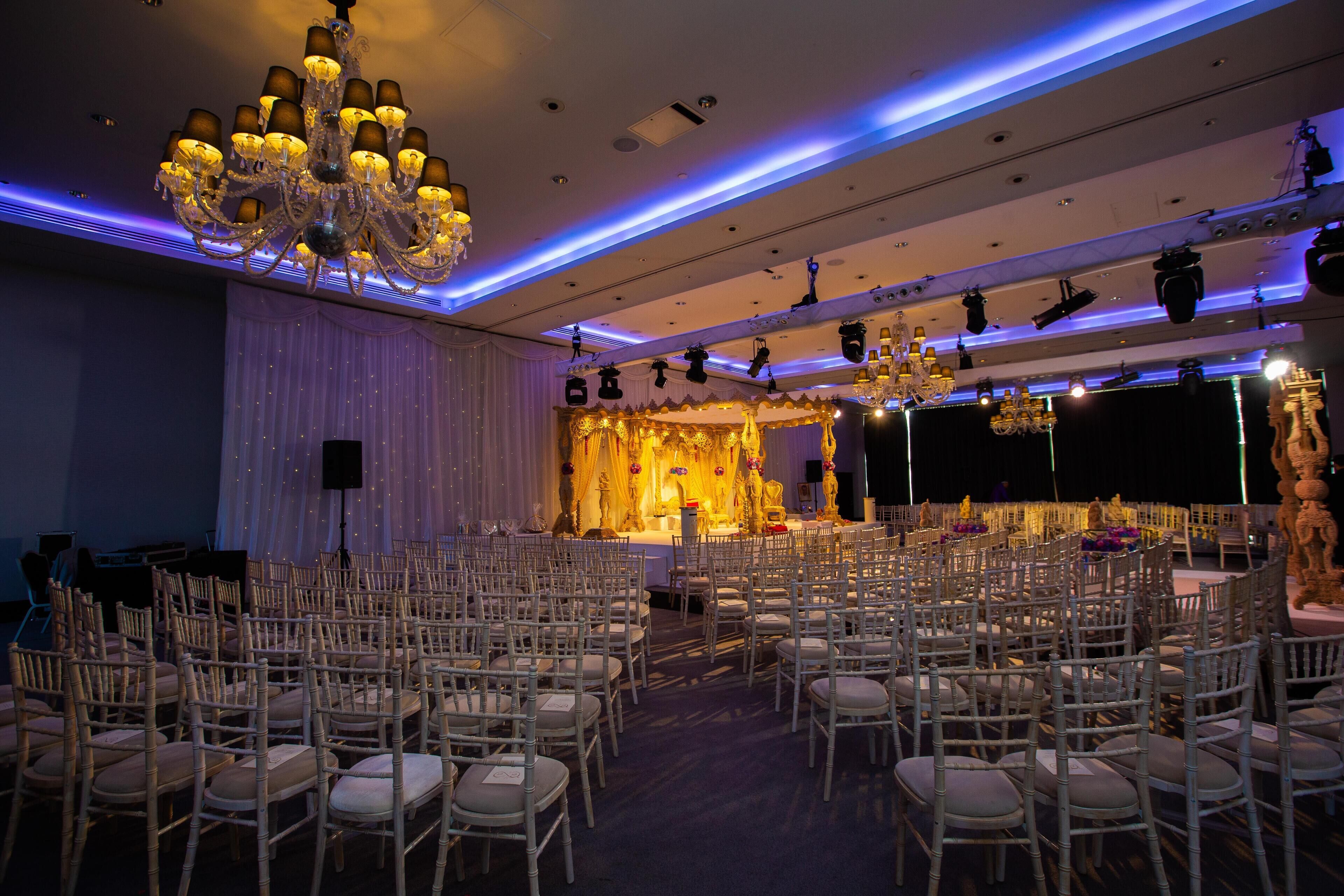 A banquet hall with rows of white chairs, a stage with a golden backdrop, and a chandelier.