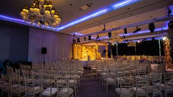 A banquet hall with rows of white chairs, a stage with a golden backdrop, and a chandelier.