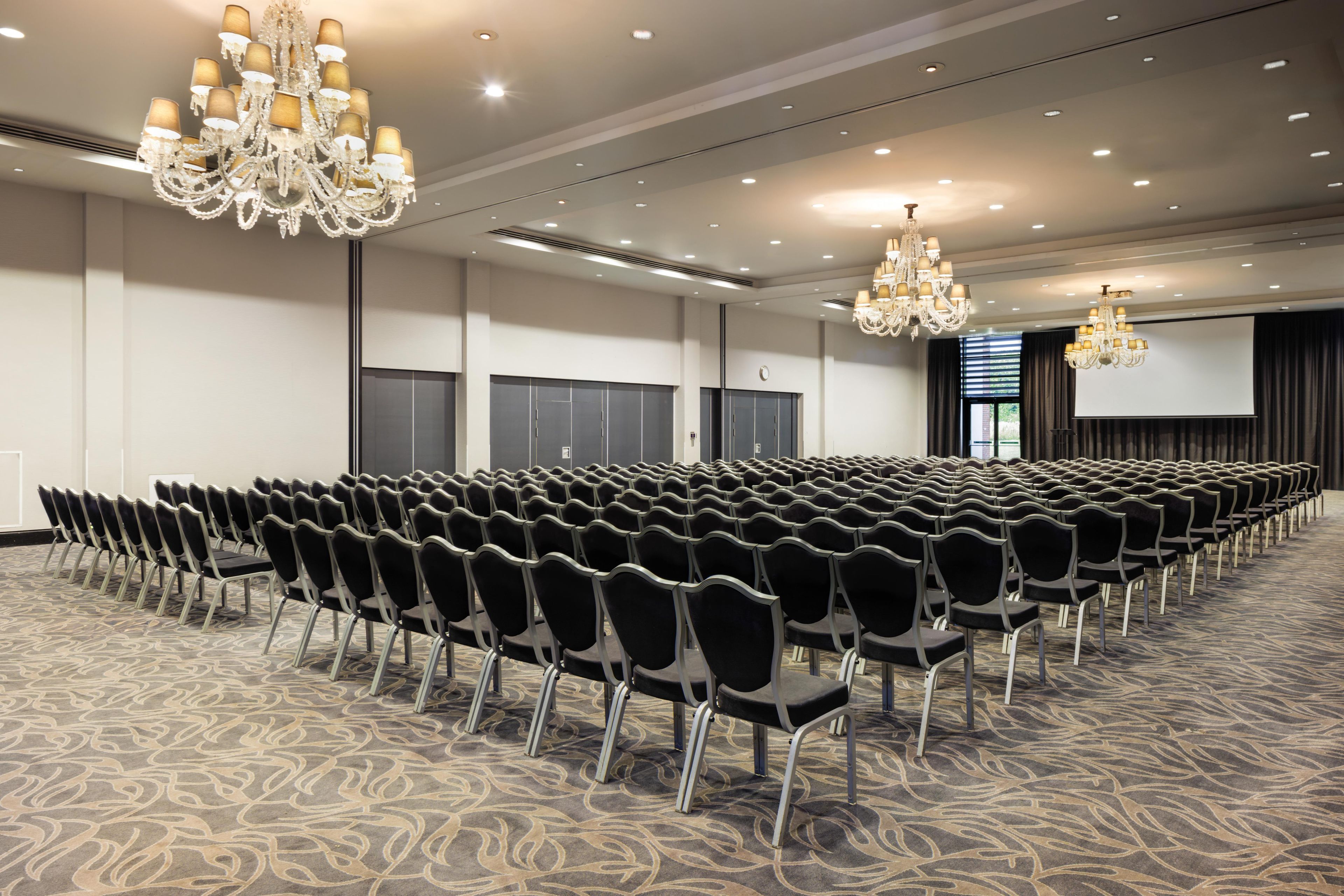 A conference room with rows of chairs, chandeliers, a large screen, and a carpeted floor.
