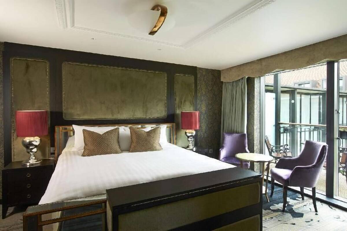 Quarto de hotel com uma cama grande, duas luminárias de cabeceira, uma mesinha e cadeiras roxas.