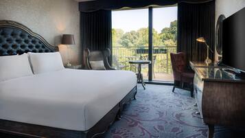 A hotel room with a large bed, a desk, a chair, and a balcony with a view of trees.