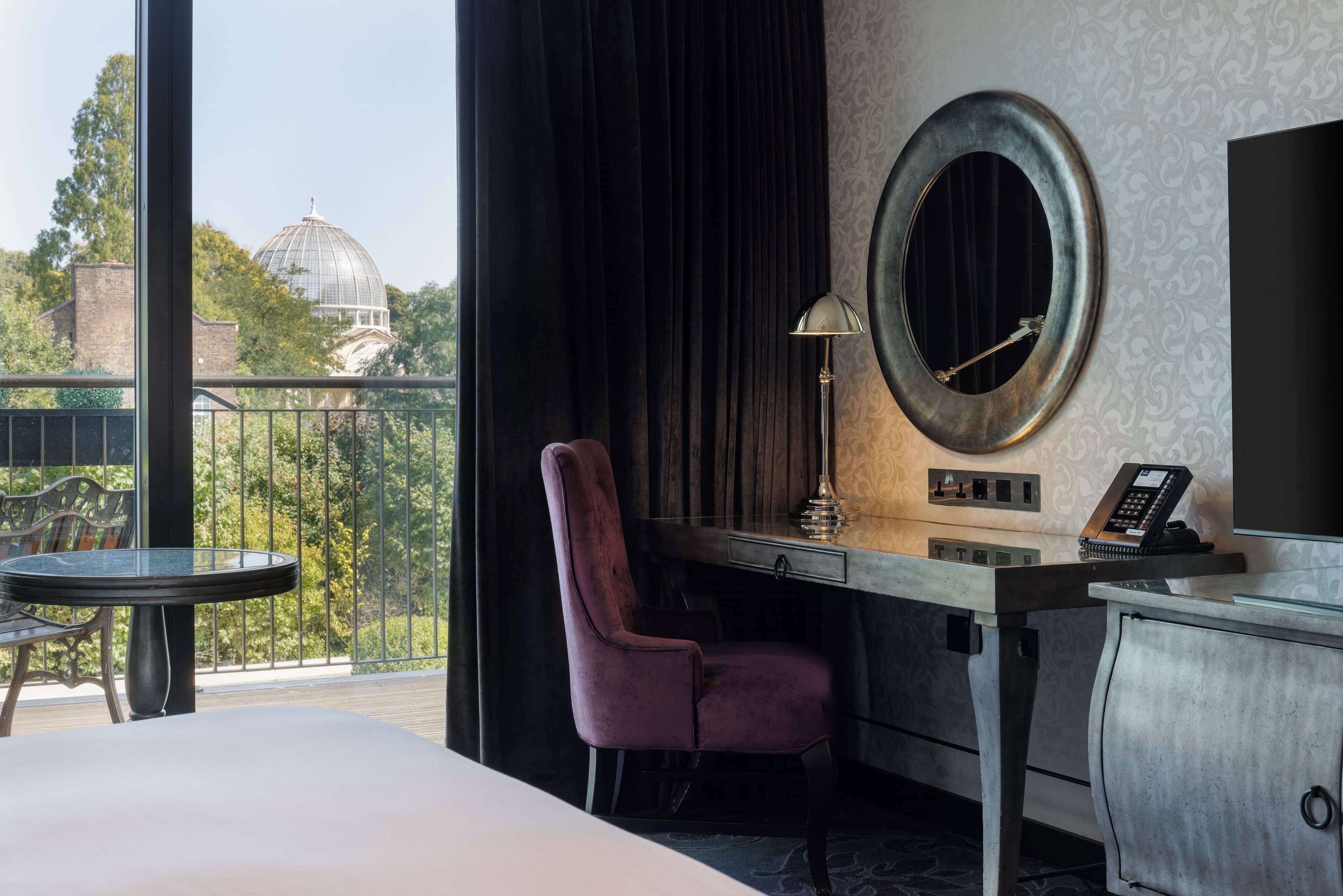 A hotel room with a large bed, a desk with a chair, a round mirror, a telephone, and a view of a dome and trees through a balcony.