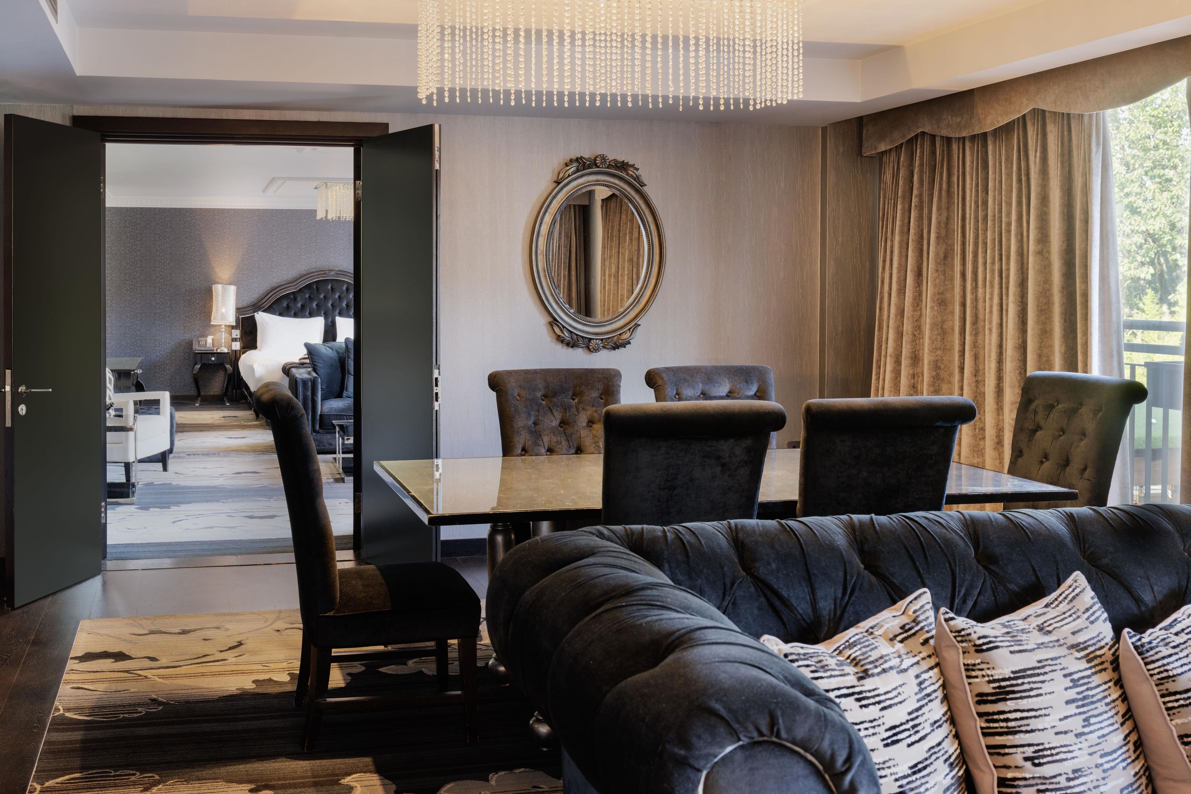 A modern living room with a black leather sofa, a dining table with chairs, a large mirror, and a view of a balcony.