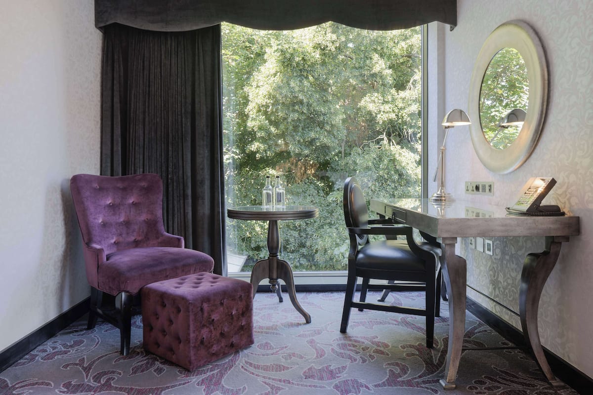 Quarto com poltrona roxa, um banquinho combinando, uma pequena mesa redonda, escrivaninha com cadeira, espelho e vista para árvores pela janela.