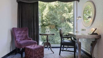 A room with a purple armchair, a matching ottoman, a small round table, a desk with a chair, a mirror, and a view of trees through a window.
