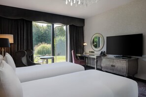 A hotel room with a large bed, a desk with a chair, a television, a mirror, and a view of the outdoors. - Hilton London Syon Park (Brentford)