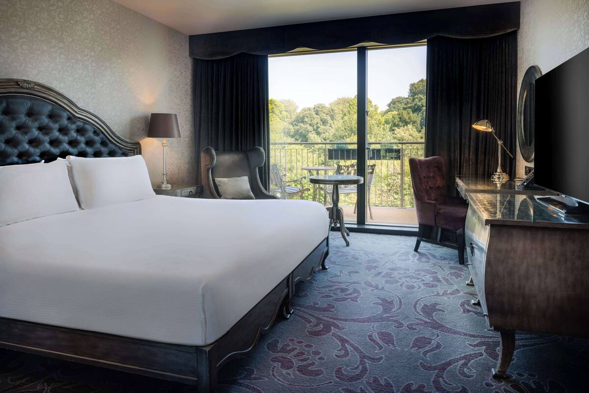 Quarto de hotel com uma cama grande, uma escrivaninha, uma cadeira, uma mesinha e uma varanda com vista para árvores.
