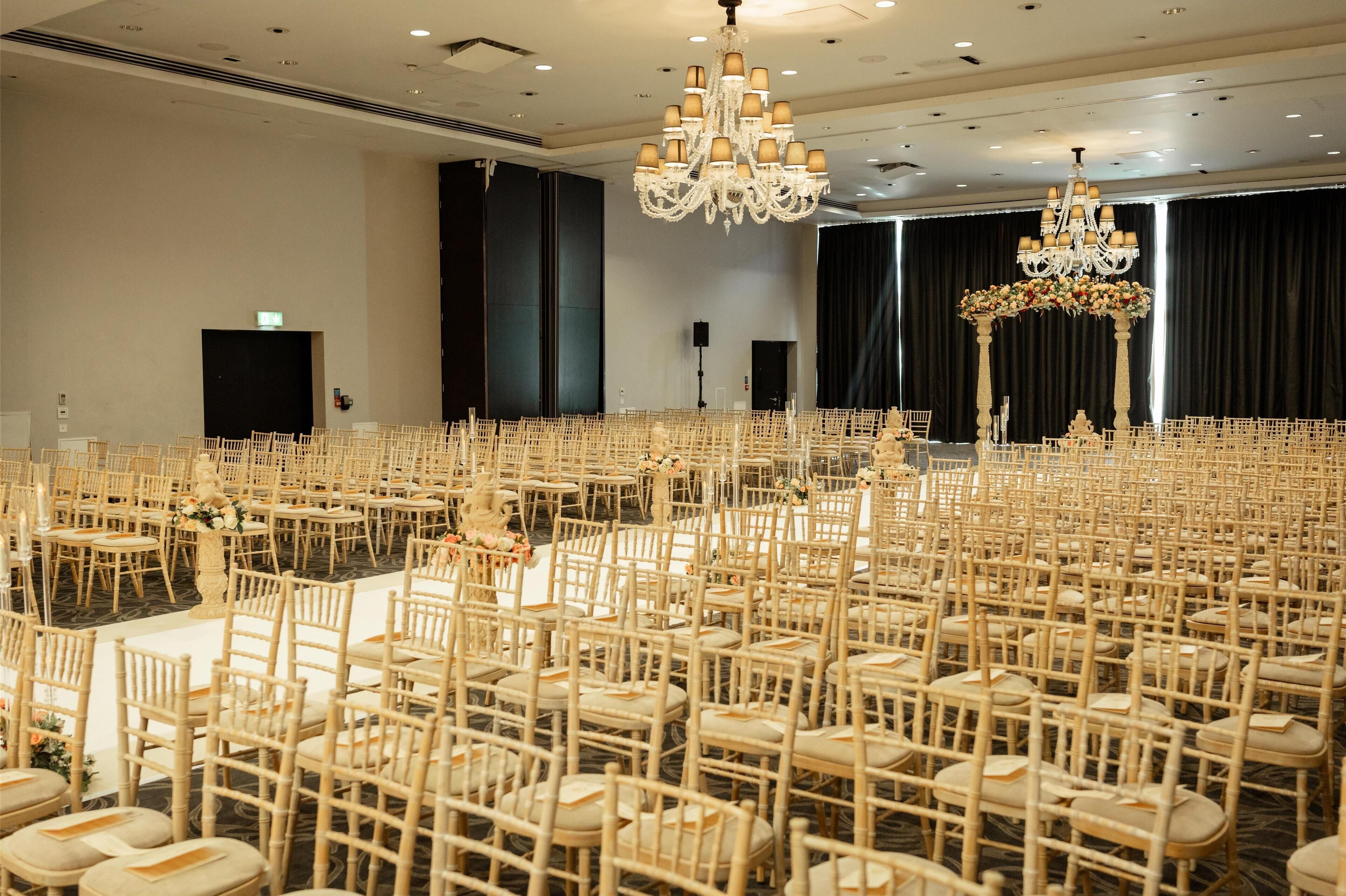 A large banquet hall with rows of wooden chairs and tables, chandeliers, and a stage area.