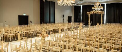 A large banquet hall with rows of wooden chairs and tables, chandeliers, and a stage area.