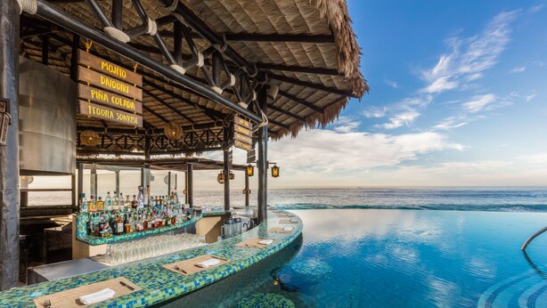 Una piscina infinita con zona de bar que presenta una variedad de botellas y una estructura con techo de paja.