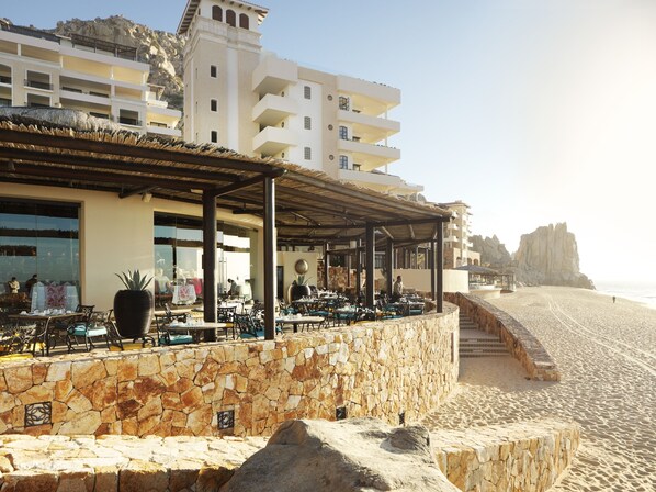 Un restaurante frente al mar con terraza, paredes de piedra y vistas a la playa y acantilados rocosos.