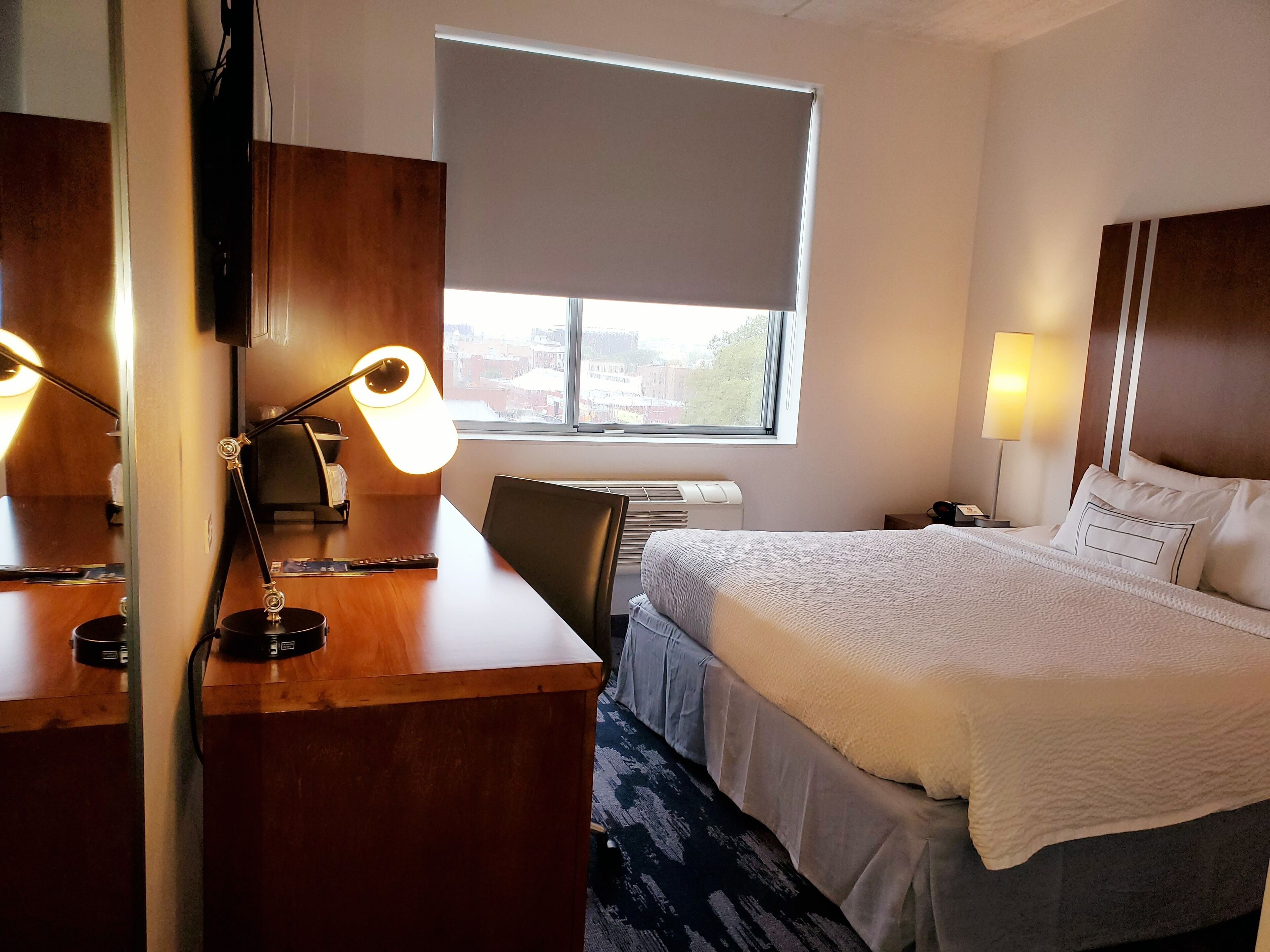 A hotel room with a bed, a desk with a lamp, a chair, a nightstand, and a window with blinds.