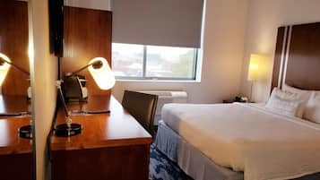 A hotel room with a bed, a desk with a lamp, a chair, a nightstand, and a window with blinds.