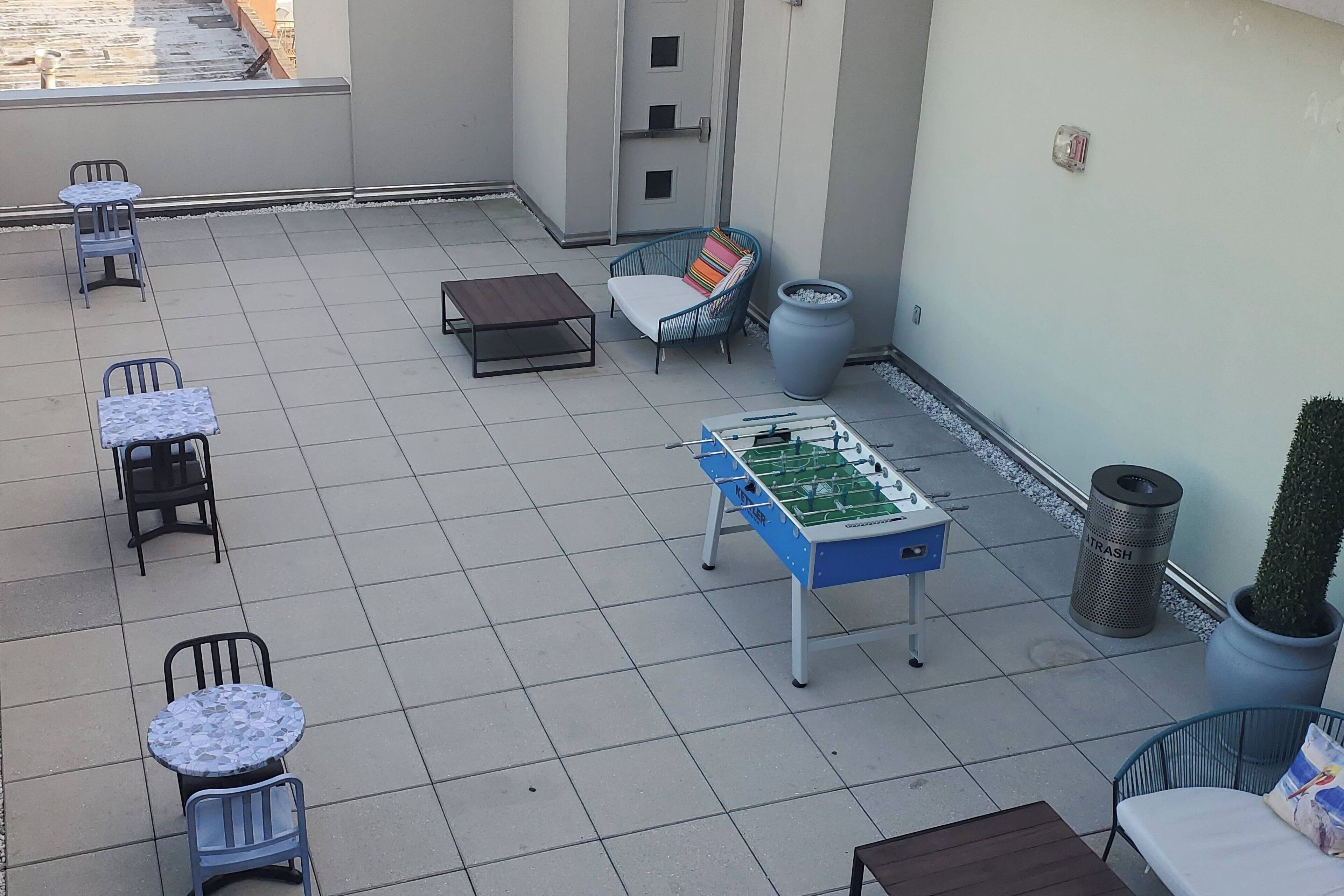 Una terraza con mesas y sillas, una mesa de fútbol de mesa, un cubo de basura y una planta en maceta.