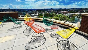 Een terras op het dak met kleurrijke stoelen en tafels van draad, met uitzicht op een stadsbeeld.