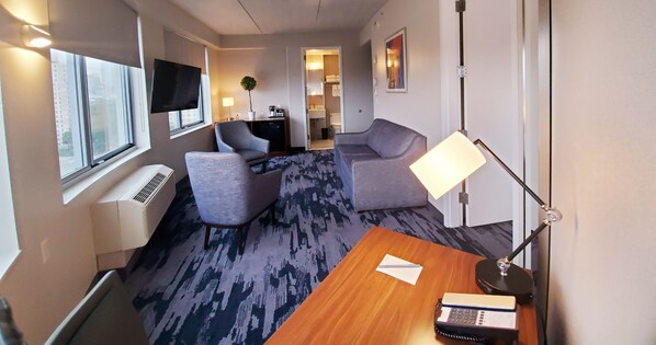 Habitación de hotel con un ventanal grande, un escritorio con lámpara, un sofá y un televisor en la pared.