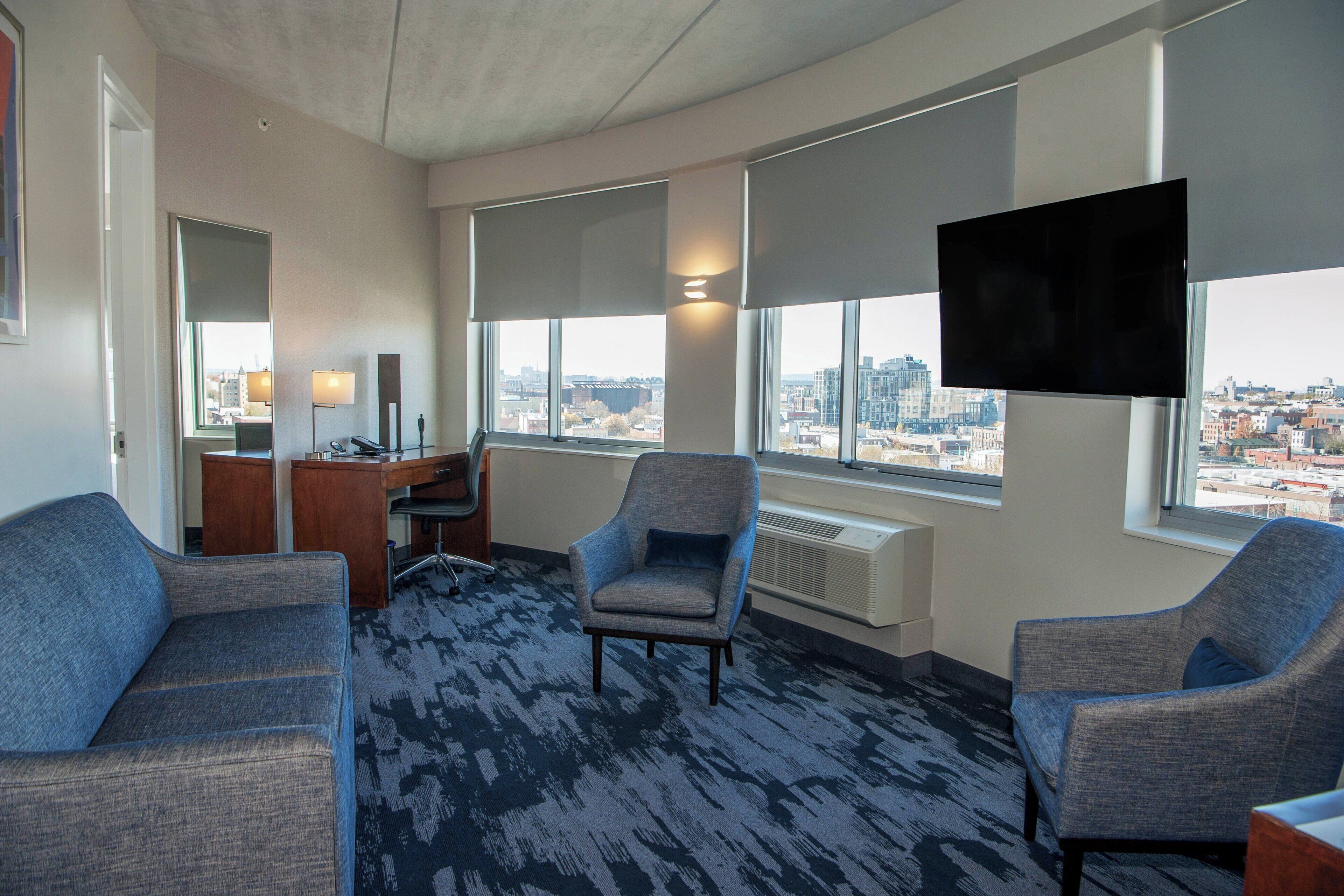 Hotelkamer met uitzicht over de stad, een bureau met lamp, twee fauteuils en een flatscreen televisie.