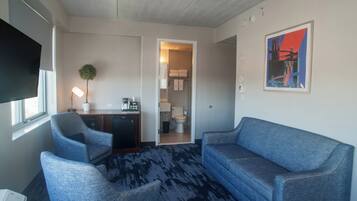 Ein Hotelzimmer mit einem großen Flachbildfernseher, zwei Sesseln, einem Sofa, einem kleinen Schreibtisch mit Lampe und einem gerahmten Bild an der Wand.