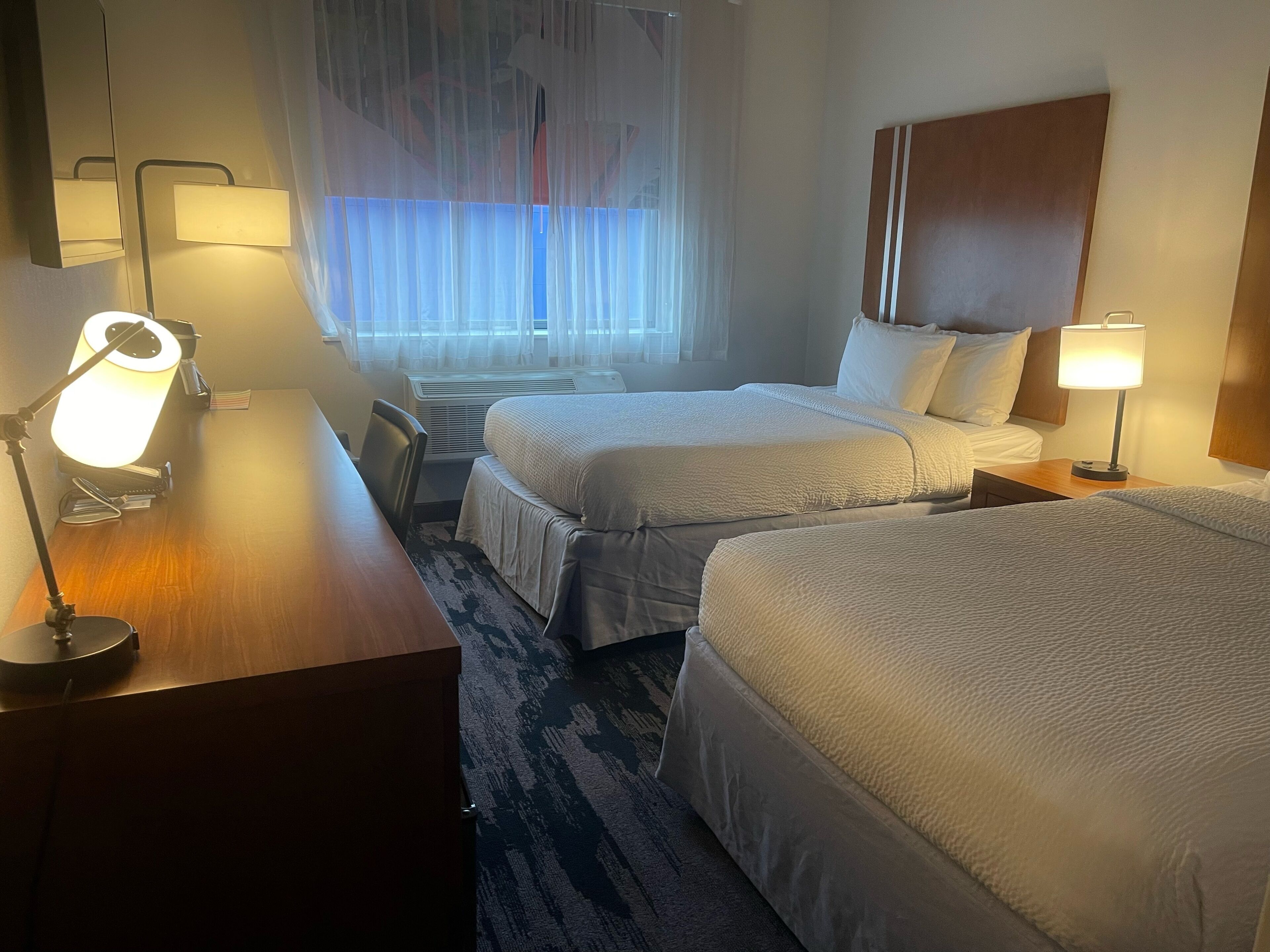 Habitació d'hotel amb dos llits, un escriptori amb una làmpada, una cadira i una finestra amb cortines.