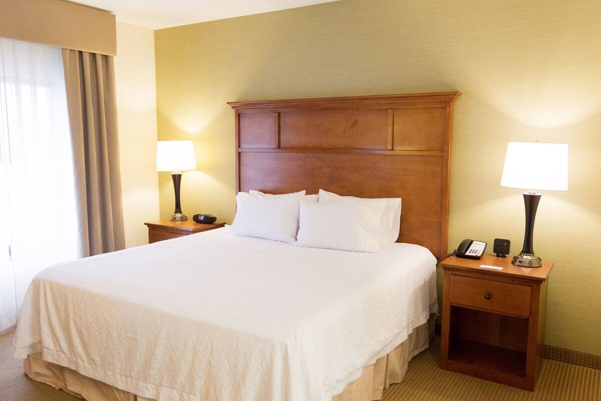 A hotel room with a large bed, two bedside tables, a nightstand, a lamp, a telephone, and a window with curtains.