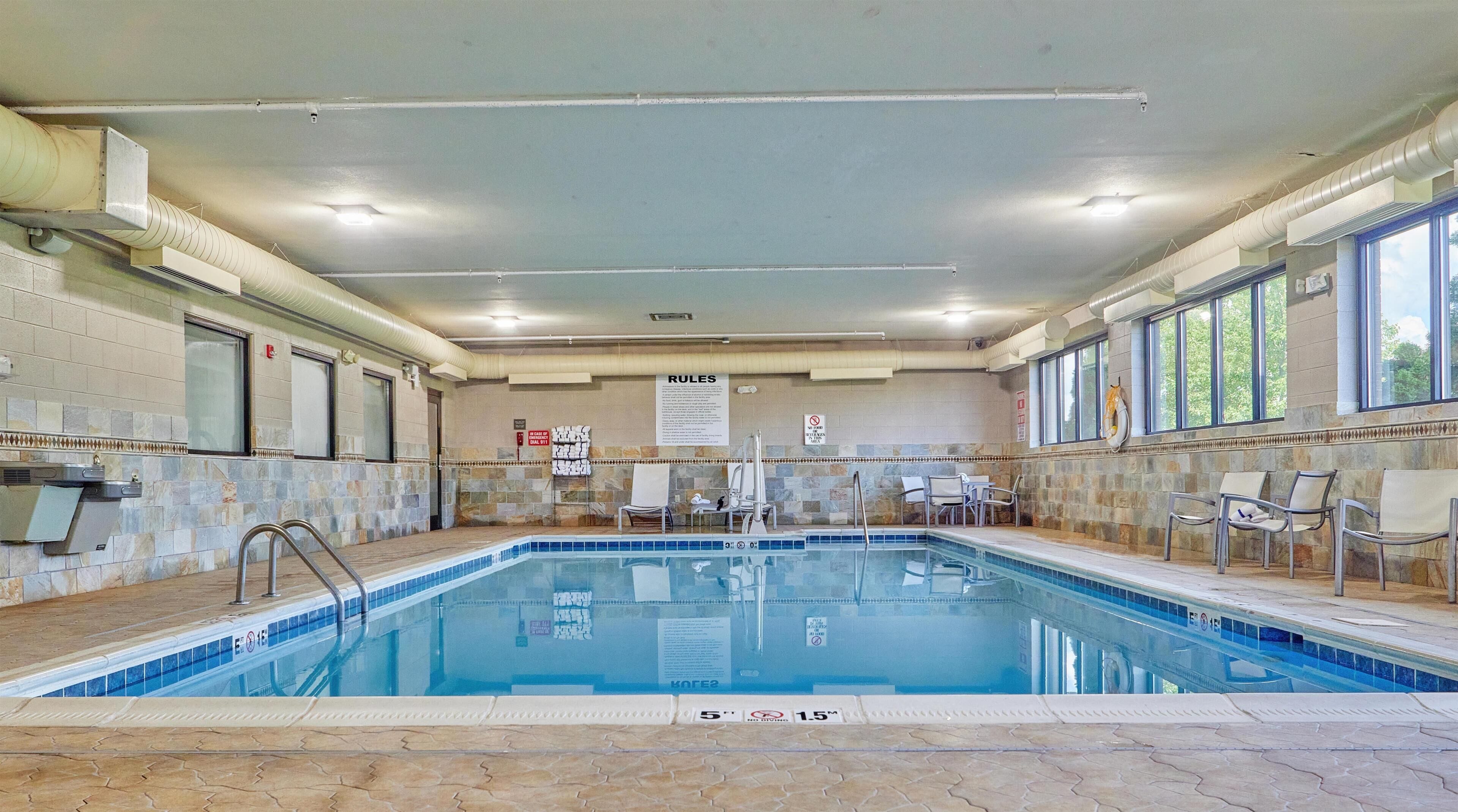 En indendørs swimmingpool med flisebeklædte vægge, et loft med ventilationskanaler og et skilt med regler.