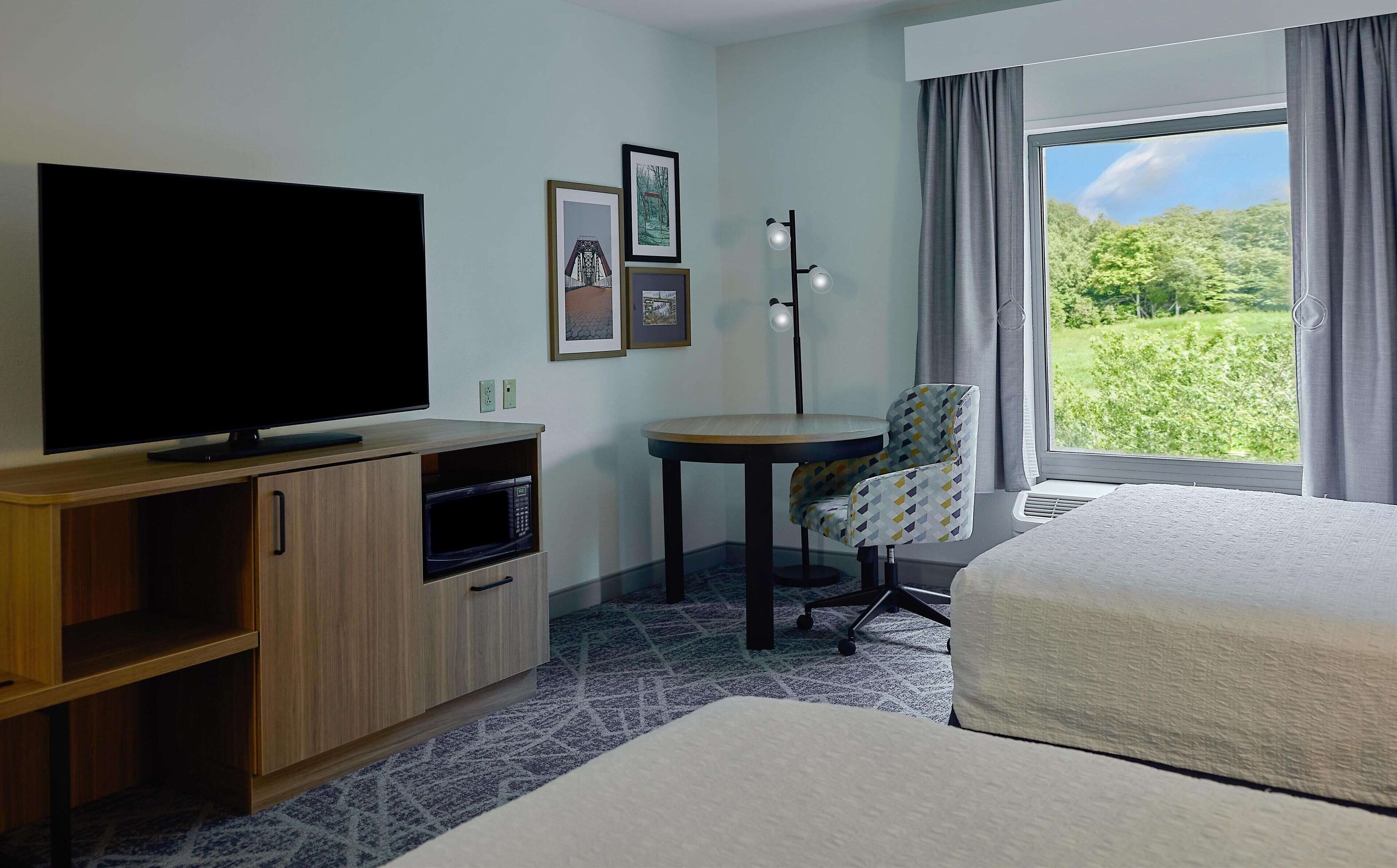 Een hotelkamer met een bed, een televisie op een houten kast, een kleine tafel met een stoel, en een groot raam met uitzicht op groen.