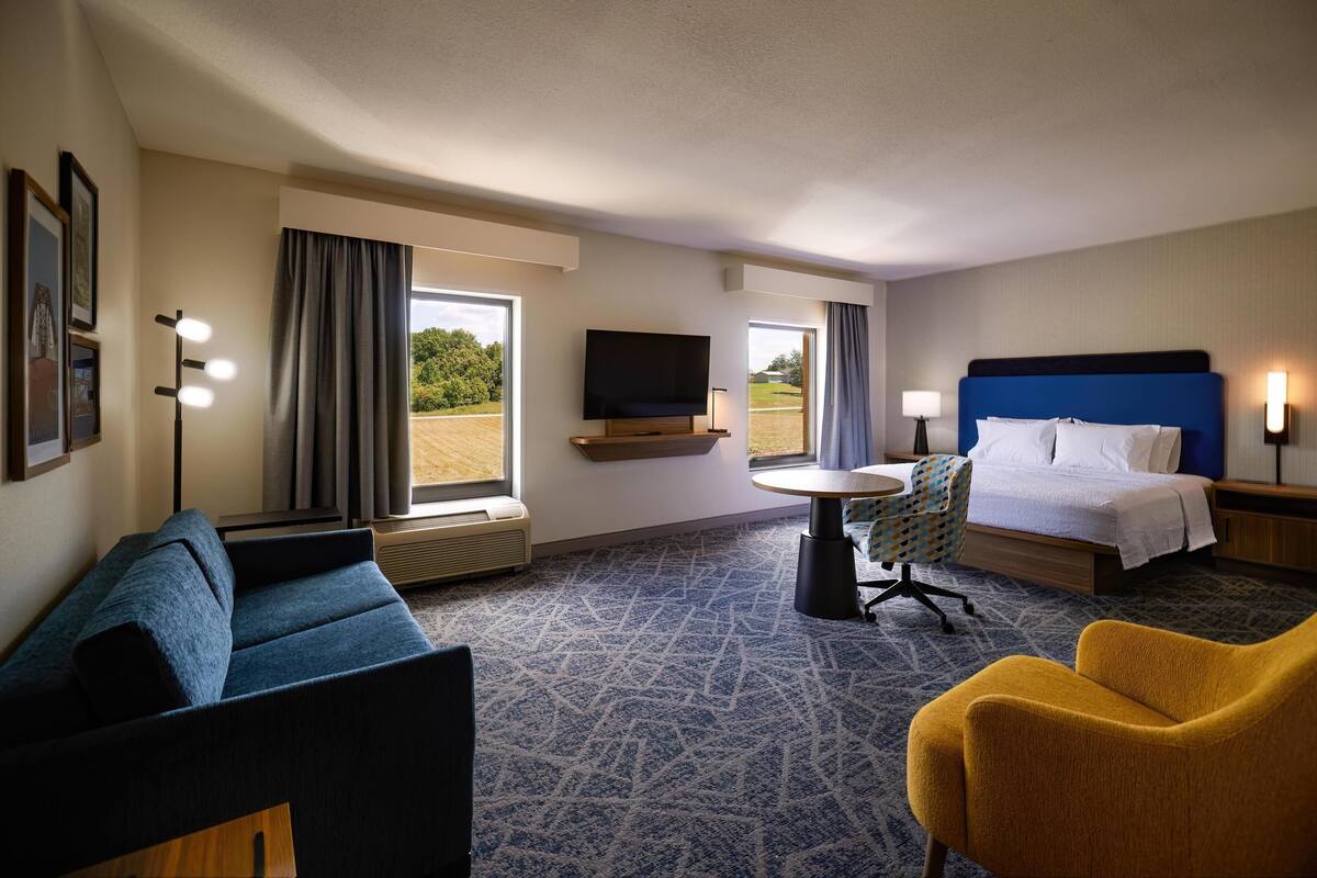 A hotel room with a bed, a sofa, a chair, a small table, a TV, and large windows with curtains.
