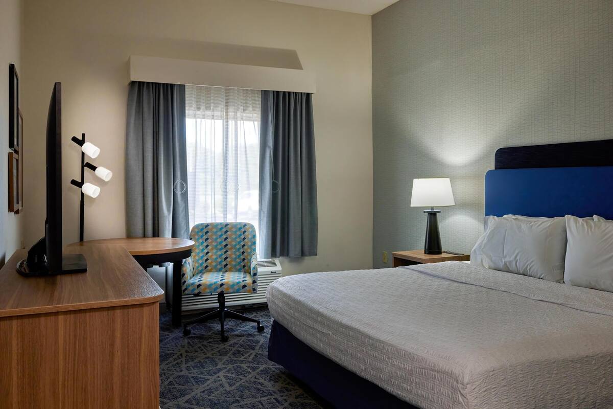 A hotel room with a bed, a desk, a chair, a lamp, and a window with curtains.
